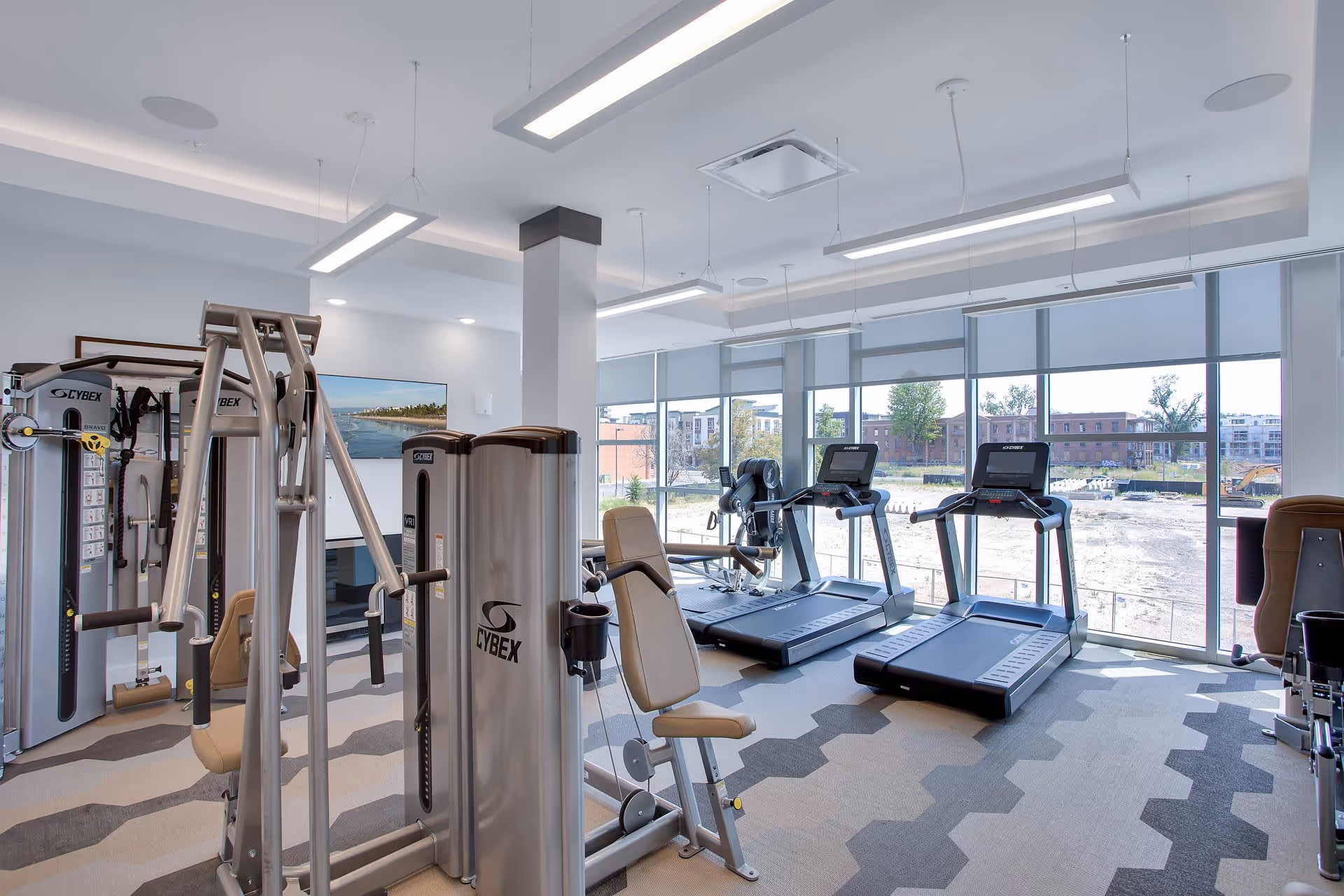A modern fitness center with various exercise machines including treadmills, weight machines, and a stationary bike. The room has large windows allowing natural light and a view of buildings and trees outside. The floor has a geometric patterned carpet and the ceiling has suspended rectangular lights.