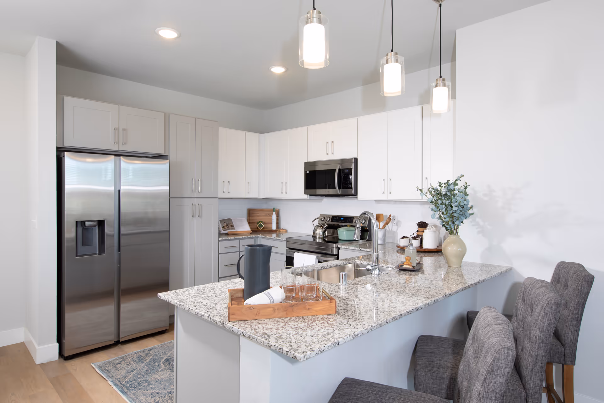 Modern kitchen with white cabinets, stainless steel refrigerator, stove, and microwave. Granite countertops with a sink and faucet, decorated with a wooden tray holding glasses and a pitcher, and a vase with greenery. Three pendant lights hang from the ceiling, and two gray upholstered chairs are positioned at the counter.
