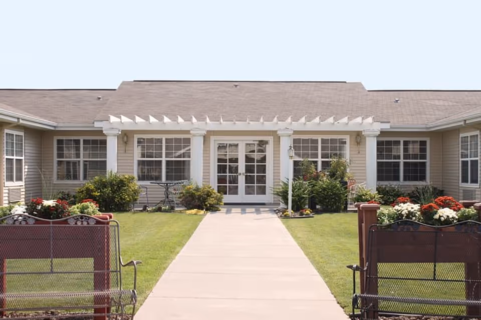 Front exterior view of a single-story senior living facility building with beige siding, white trim, and a pergola above the entrance. There is a concrete walkway leading to double glass doors, flanked by windows and landscaping with green bushes and colorful flowers. Two metal benches are positioned on either side of the walkway.