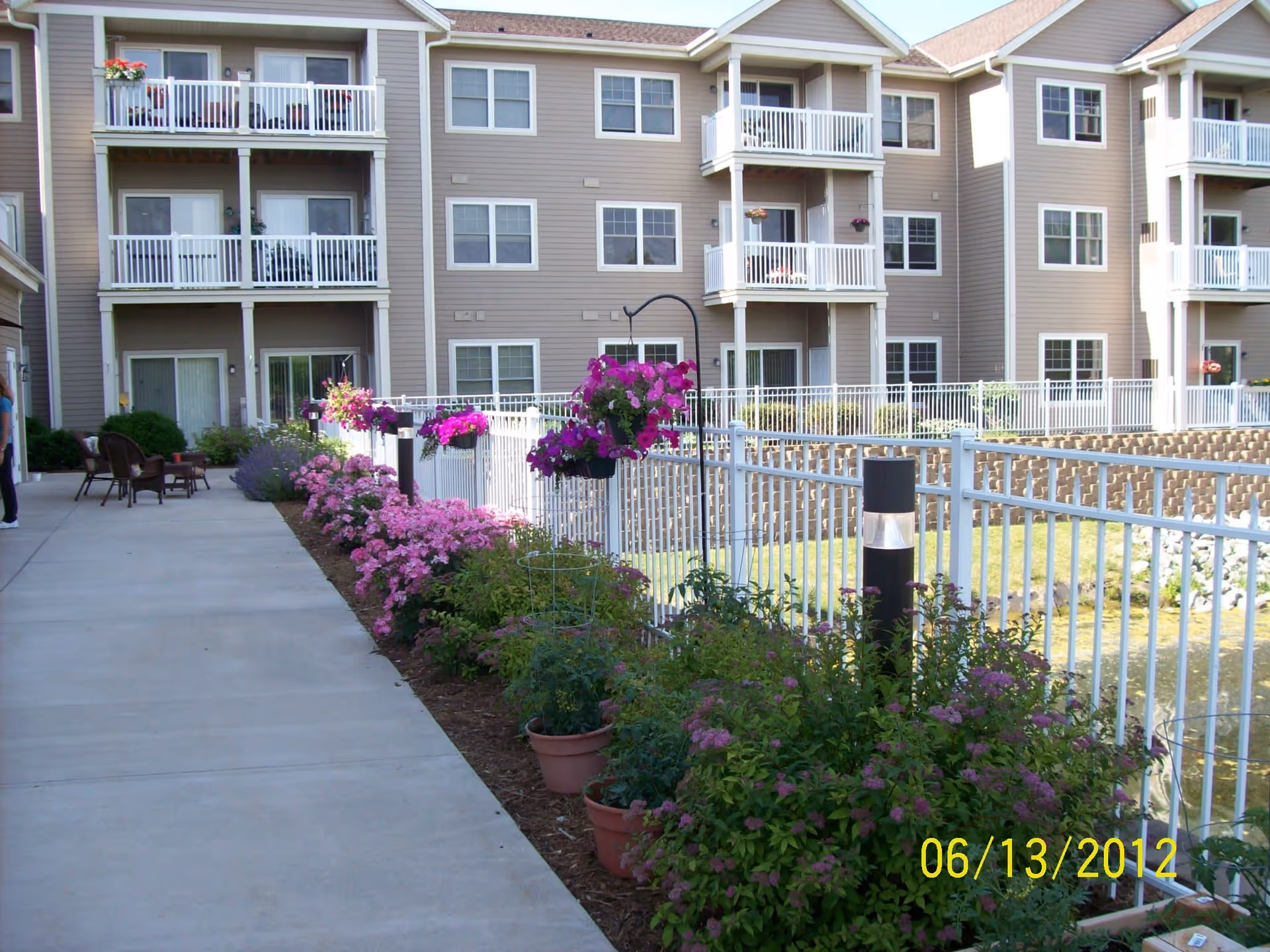 Outdoor walkway beside a multi-story residential building with balconies. The walkway is lined with flower beds containing pink and purple flowers, hanging flower baskets, and potted plants. There is a white metal fence separating the walkway from a grassy area with a retaining wall. A person is partially visible walking on the left side of the walkway.