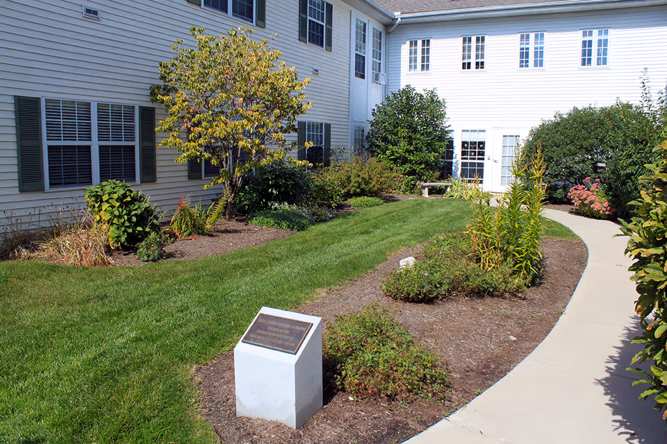 A landscaped outdoor garden area at Ohio Living Swan Creek with a curved concrete pathway, green grass, various shrubs, and small trees. The garden is bordered by a white building with multiple windows and green shutters. A small stone monument with a plaque is visible in the foreground.