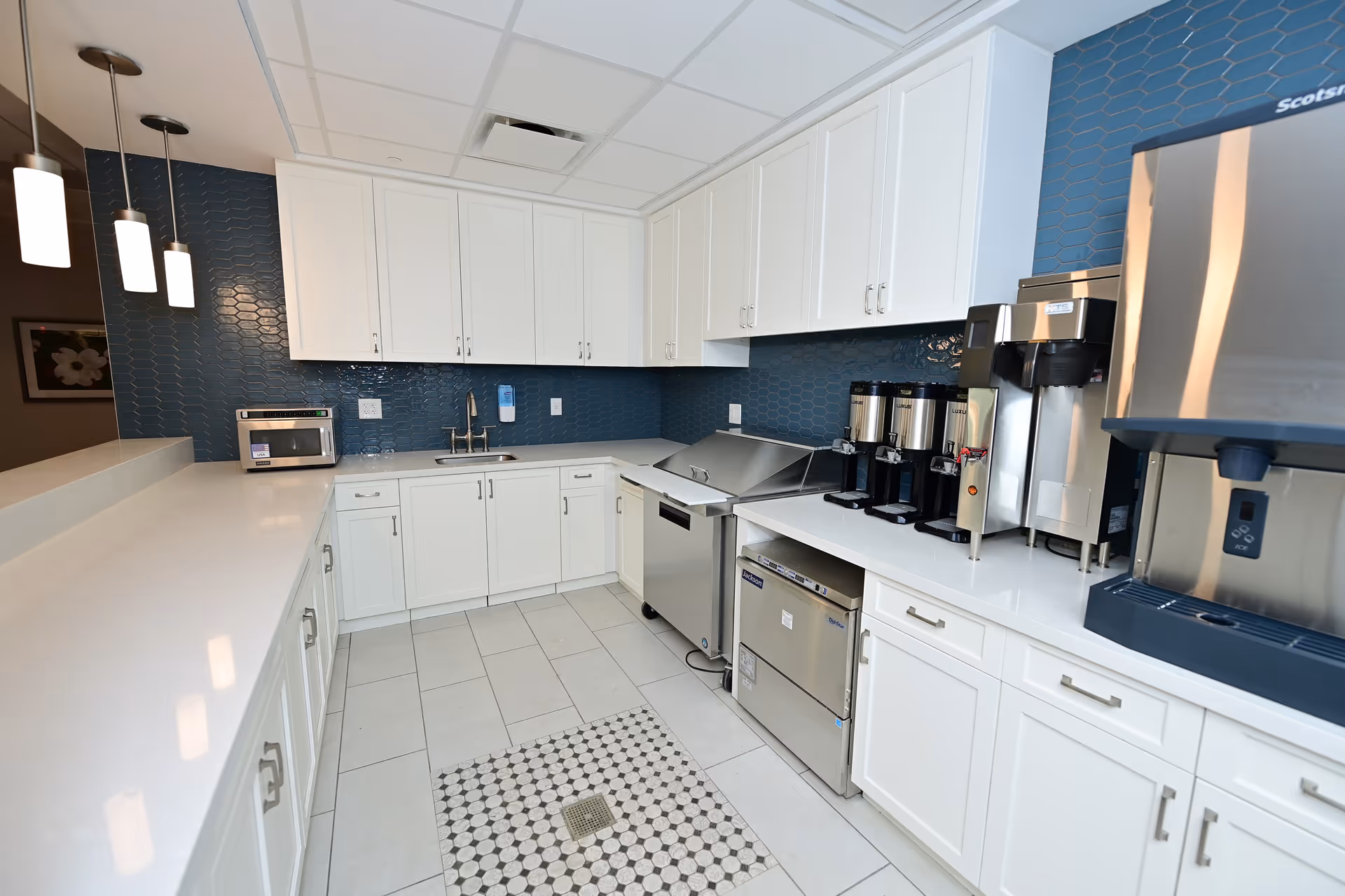 A modern kitchen area with white cabinets and countertops, blue hexagonal tile backsplash, stainless steel appliances including coffee machines and a water dispenser, and three pendant lights hanging over a counter.