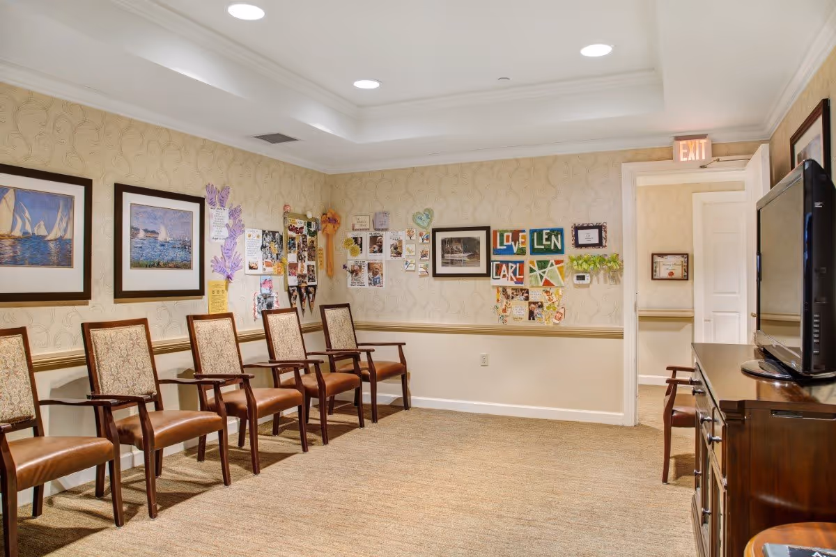 A waiting or common area in a senior living facility with a row of six wooden chairs with cushioned seats and backs along one wall. The walls are decorated with framed pictures of sailboats and a bulletin board filled with colorful artwork, notes, and photos. There is a wooden cabinet with a flat-screen TV on top on the right side of the room. The room has beige patterned wallpaper, carpeted floor, and recessed ceiling lights. An exit sign is visible above an open door leading to another room.