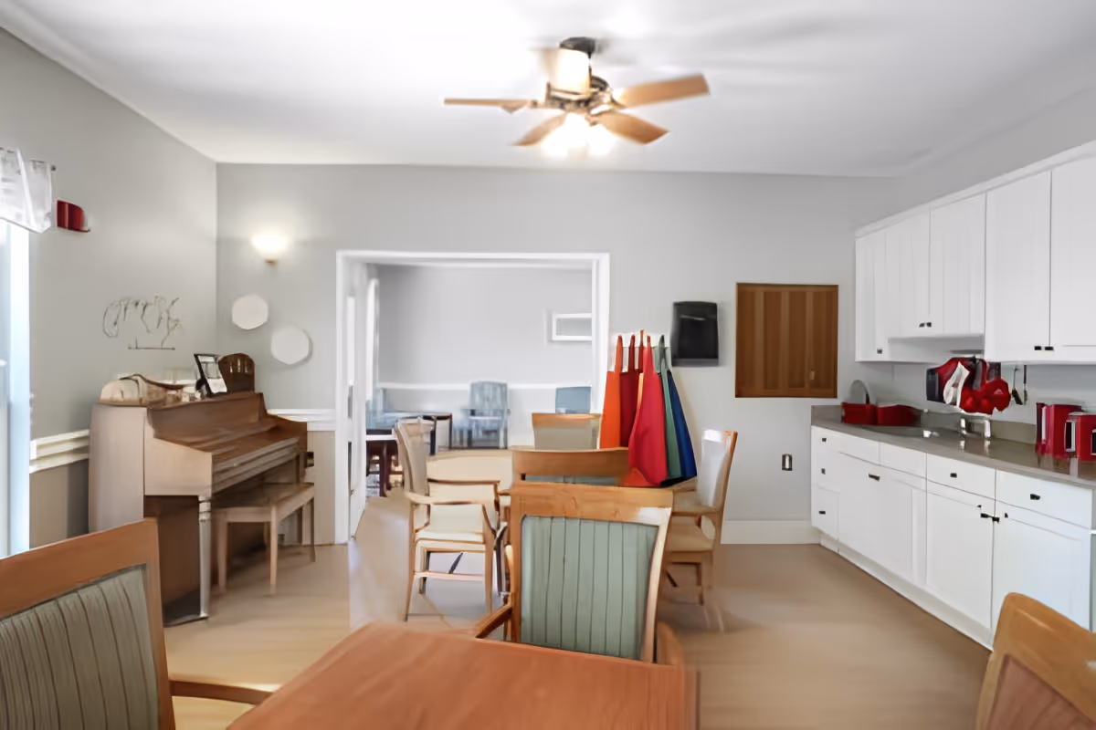 A bright room with wooden tables and chairs, a piano against the left wall, and a ceiling fan with lights. The room has white cabinets and a countertop on the right side with red kitchen appliances and utensils. Colorful aprons hang on a rack near the center, and another room with additional seating is visible through an open doorway.