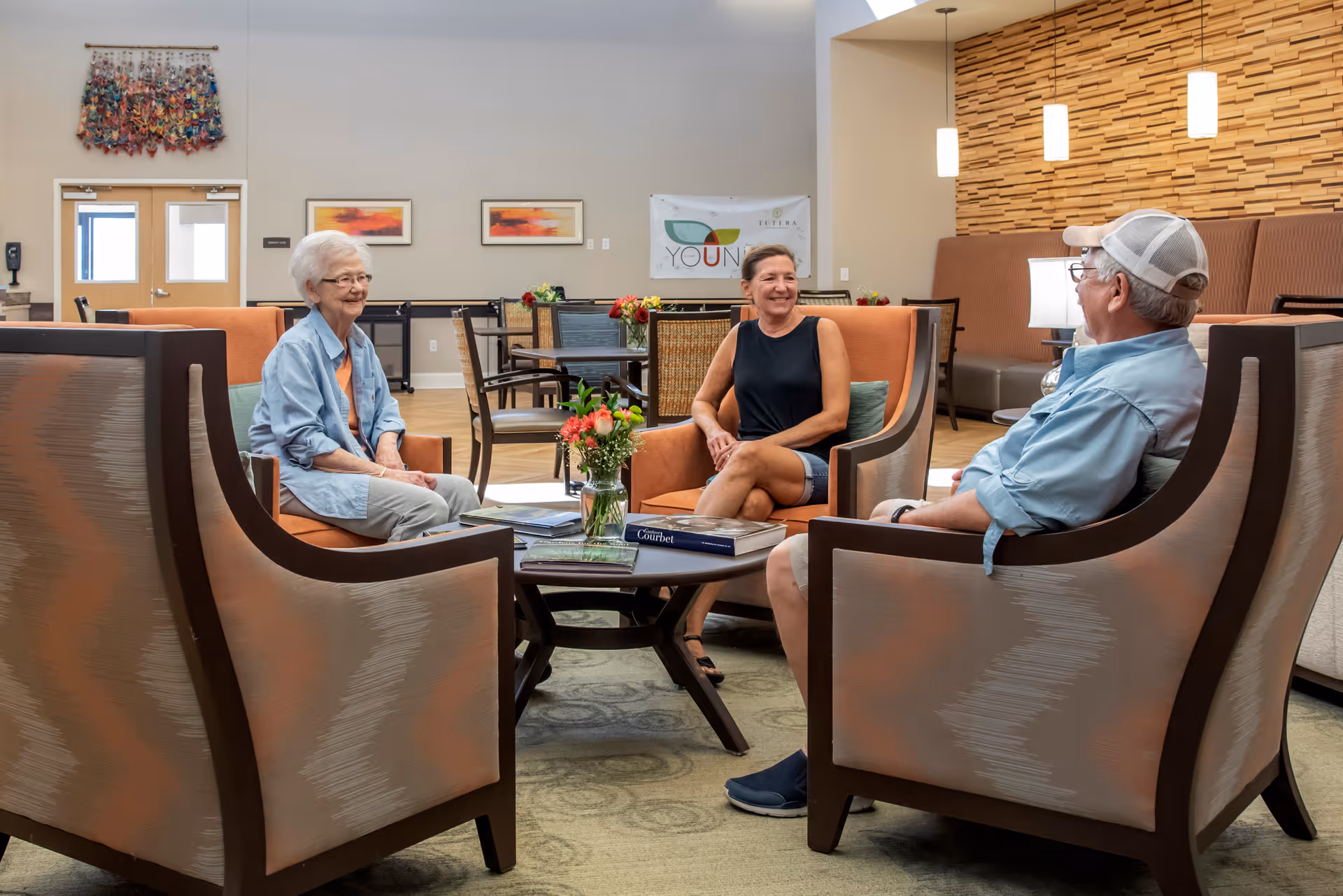 Three people sitting and chatting in a comfortable, well-lit common area with modern chairs and a coffee table with books and a vase of flowers. The room has a decorative wooden wall panel, hanging pendant lights, and artwork on the walls.