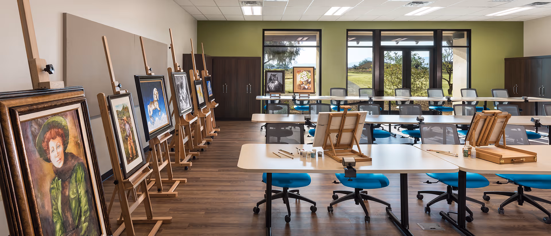 A spacious art studio room with wooden easels displaying various paintings along the left wall. Several tables with blue swivel chairs are arranged in the center and back of the room, each table equipped with art supplies such as paintbrushes and paint bottles. Large windows at the back let in natural light and offer a view of greenery outside. The room has a wooden floor and light green accent walls.