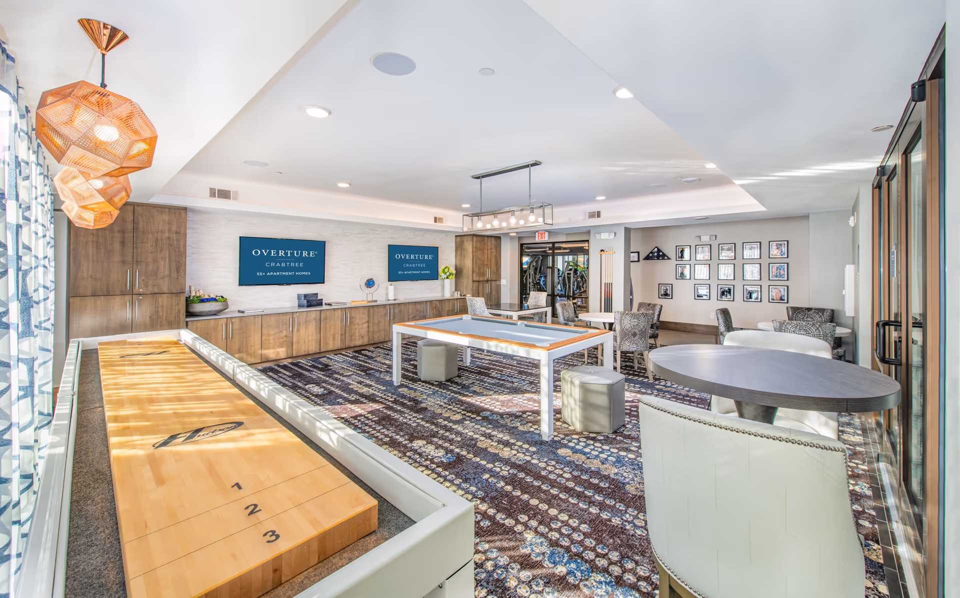 A bright and spacious recreational room in Overture Crabtree 55+ Apartment Homes featuring a shuffleboard table on the left, a pool table in the center, several round tables with chairs, patterned carpet, modern light fixtures, and two wall-mounted screens displaying the facility name.