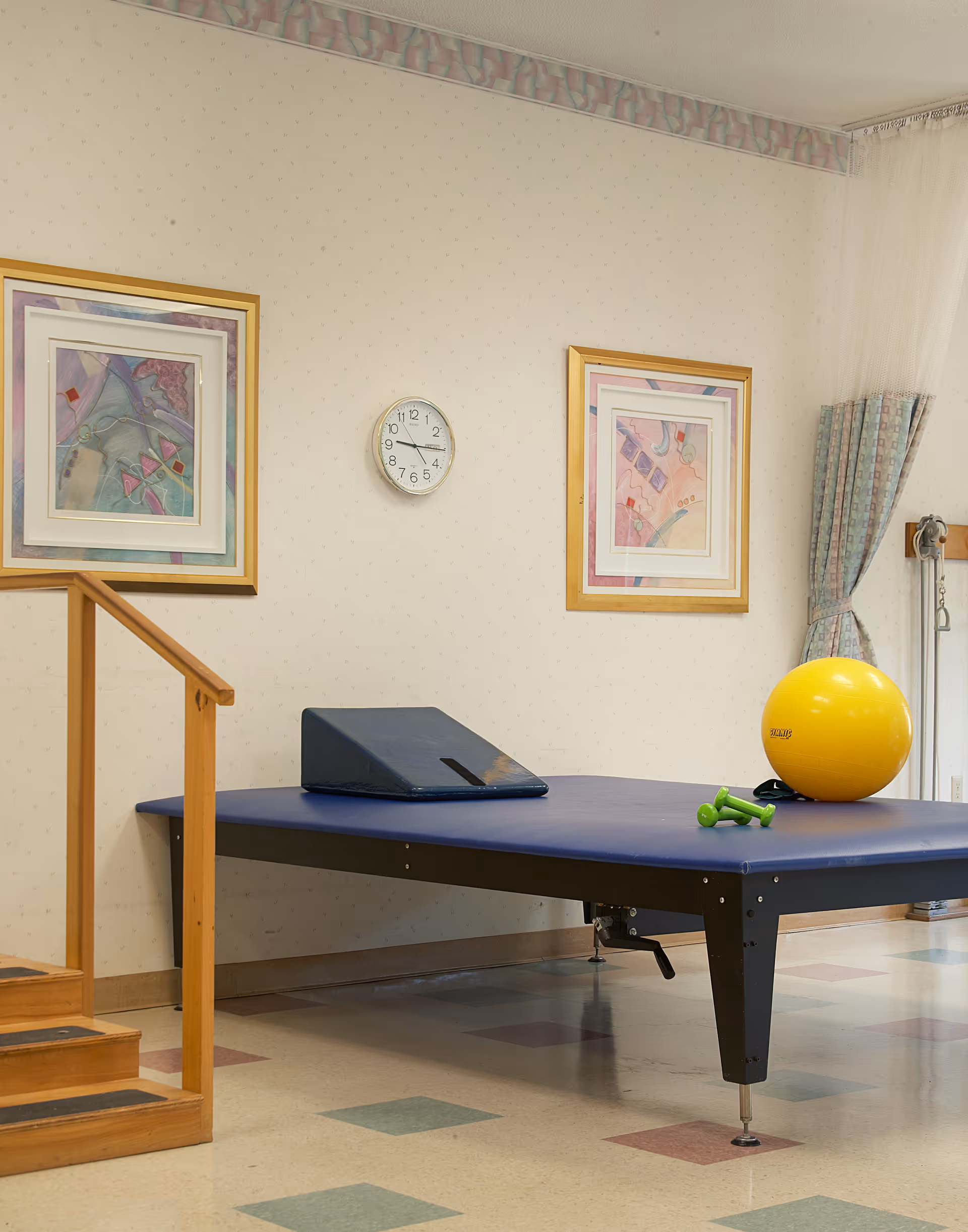 Physical therapy room with a blue padded therapy table, a yellow exercise ball, small green dumbbells, wooden handrails on stairs, two framed abstract paintings on the wall, a clock showing 2:15, and a window with patterned curtains.