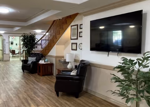 Interior view of a senior living facility showing a hallway with wooden flooring, two black leather chairs with a small wooden table between them, a large flat-screen TV mounted on the wall, a potted plant, and a staircase leading to an upper floor. In the background, there is a dining area with tables and chairs.