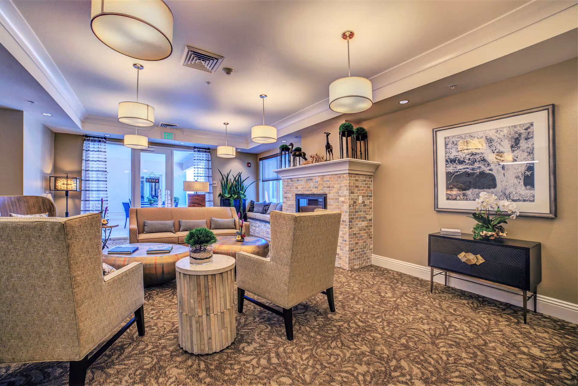 A cozy and well-lit living room area with beige armchairs, a round wooden side table, a beige sofa, and a brick fireplace adorned with decorative plants and giraffe figurines. The room has patterned carpet flooring, modern ceiling lights, a framed black and white tree artwork on the wall, and large windows with sheer curtains letting in natural light.
