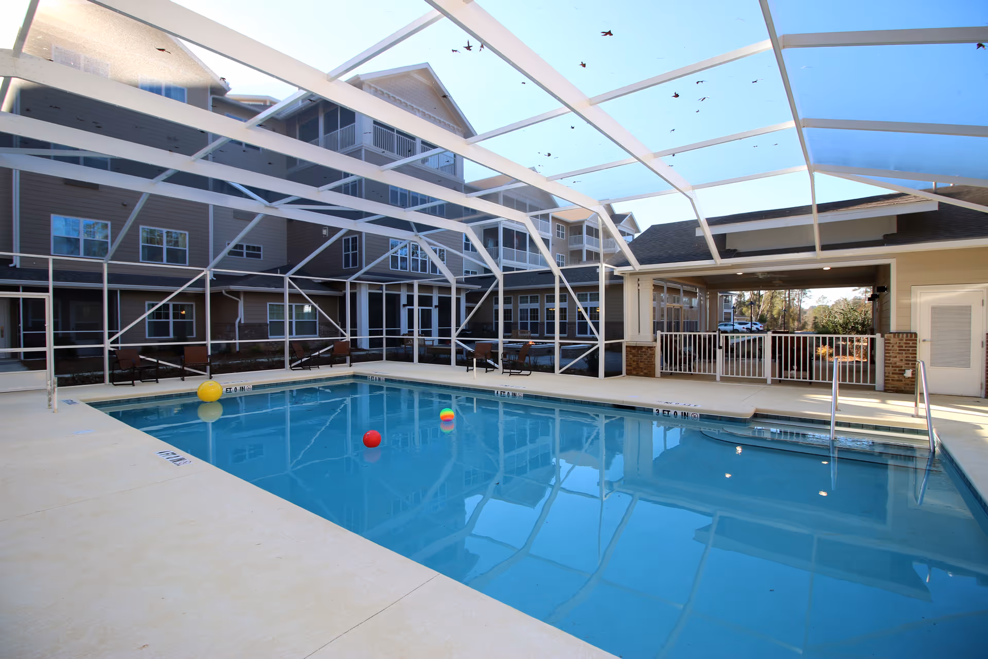 A screened outdoor swimming pool with lounge chairs and colorful floating balls beside a multi-story senior living building.