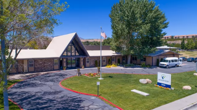Front exterior of a single-story rehabilitation center with a pitched glass entrance, circular driveway, flagpole, lawn sign reading 'ROSEWOOD', and a parked van.