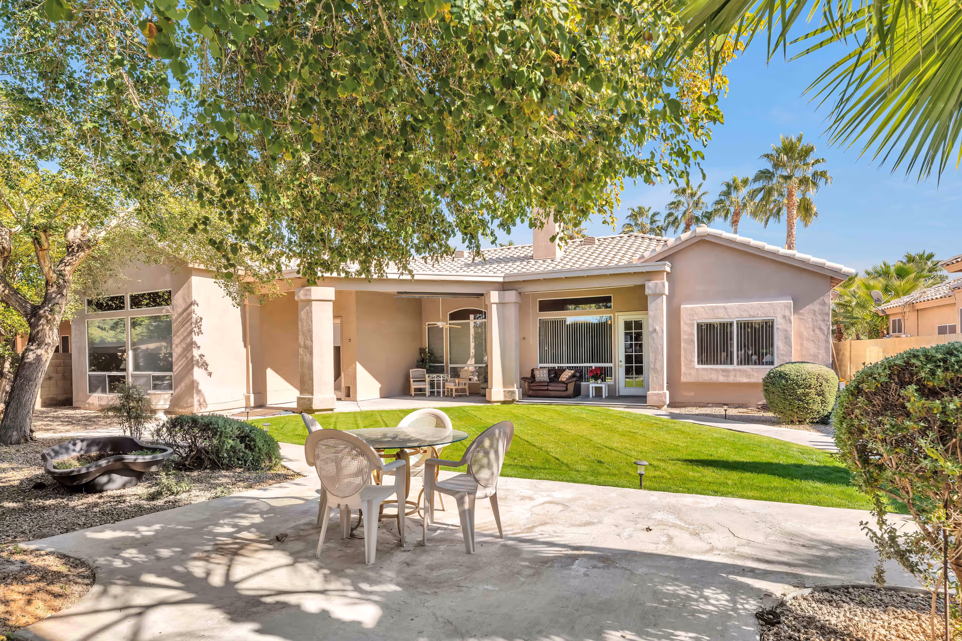 Outdoor patio area of The Sierra's Assisted Living facility with a round table and four chairs on a concrete surface, surrounded by green grass, trees, and shrubs. The building has large windows and a covered porch with seating. Palm trees are visible in the background under a clear blue sky.