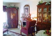 A cozy living room with a burgundy armchair, a wooden cabinet with glass doors displaying decorative items, a wooden armoire, a small fireplace with candles and framed artwork above it, a table lamp, and floral arrangements.