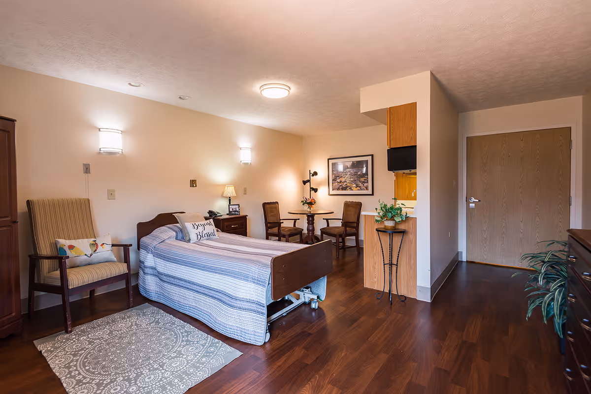 A neatly furnished senior living bedroom with a single bed, chairs, a small table, kitchenette area, and hardwood floors.
