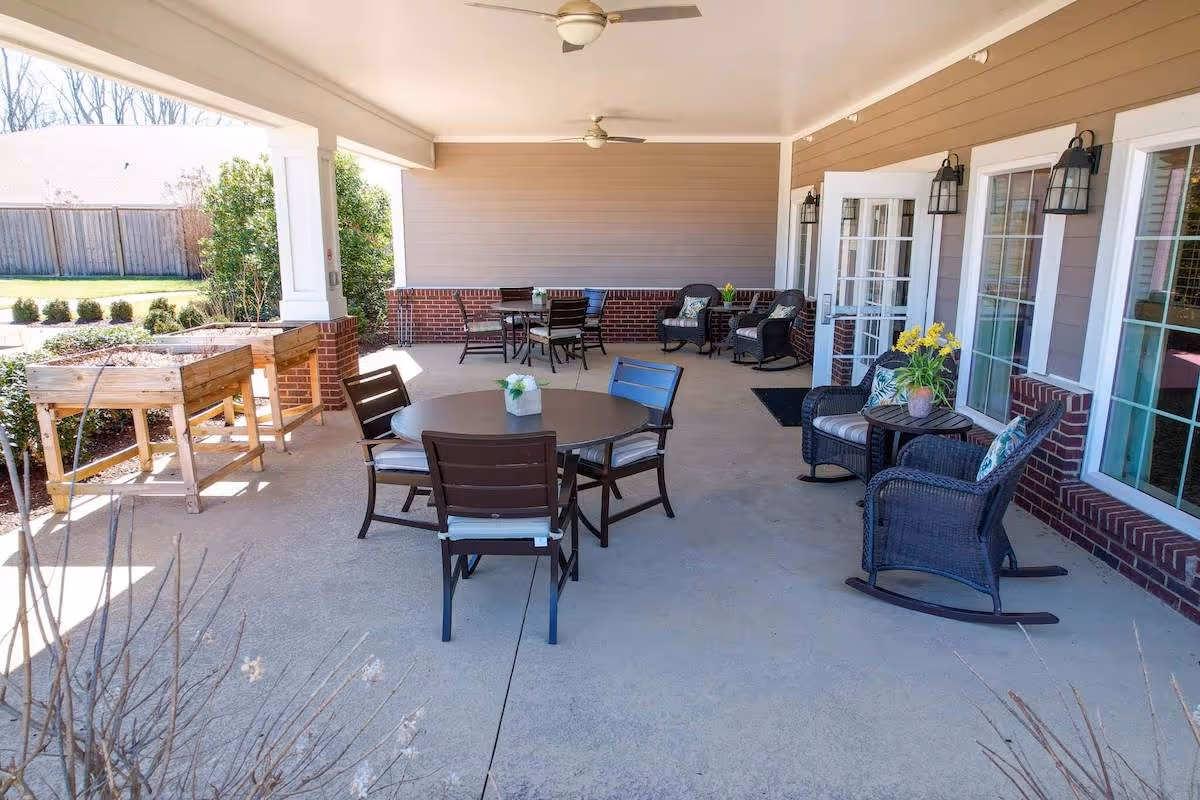 Covered outdoor patio area with several round tables and chairs, wicker rocking chairs with cushions, potted plants on tables, and raised garden beds along the left side. The patio is attached to a building with large windows and glass doors.