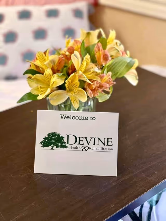 A close-up of a wooden table with a small bouquet of yellow and orange flowers in a glass vase. In front of the vase is a white card that reads 'Welcome to DEVINE Health & Rehabilitation' with a green tree logo. A blurred pillow with a pattern is visible in the background.