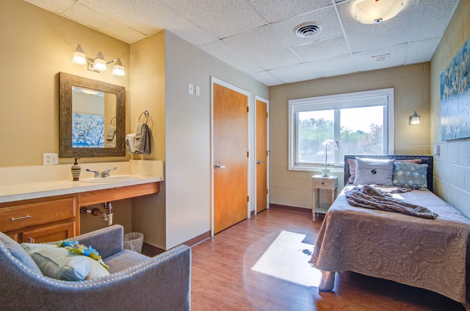 A bright and cozy senior living bedroom with a single bed covered in a patterned quilt and multiple pillows. Next to the bed is a small wooden nightstand with a white orchid plant. The room has a large window letting in natural light, a comfortable armchair with a cushion, and a vanity area with a sink, mirror, and light fixtures above. The walls are painted in neutral tones, and there are two wooden doors on one side of the room.
