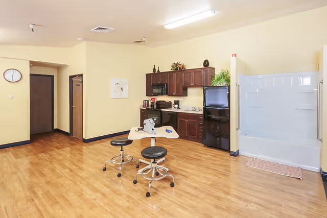 A room with light yellow walls and wooden flooring featuring a small kitchen area with dark wood cabinets, a black refrigerator, microwave, stove, and coffee maker. In the center, there is a small table with two black stools on wheels. To the right, there is a white bathtub with grab bars and a red checkered bath mat in front of it. A clock and a painting are visible on the walls.