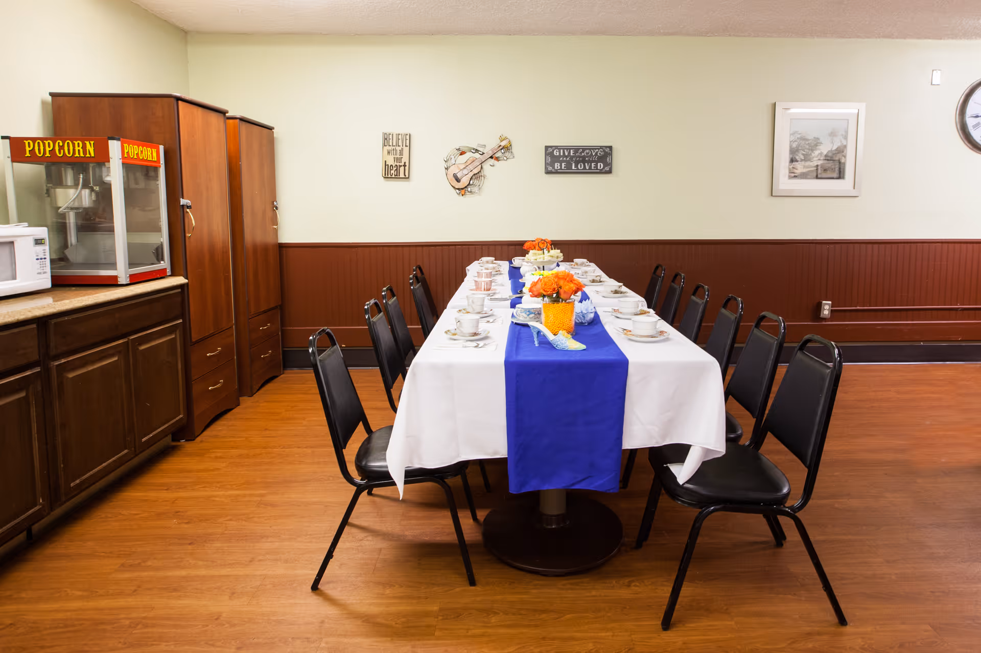 A dining room with a long table covered with a white tablecloth and a blue runner. The table is set with teacups, saucers, and plates, and decorated with orange flowers in the center. There are black chairs arranged around the table. On the left side, there is a popcorn machine and a microwave on a countertop with wooden cabinets below. The walls are light green with brown wainscoting, decorated with framed pictures and wall art including a guitar and inspirational quotes.