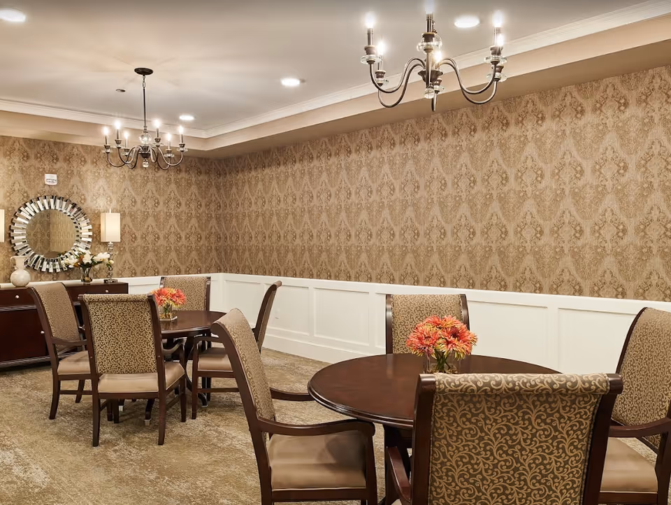 Elegant dining room with round wooden tables, upholstered chairs, chandeliers, patterned wallpaper, and floral centerpieces.