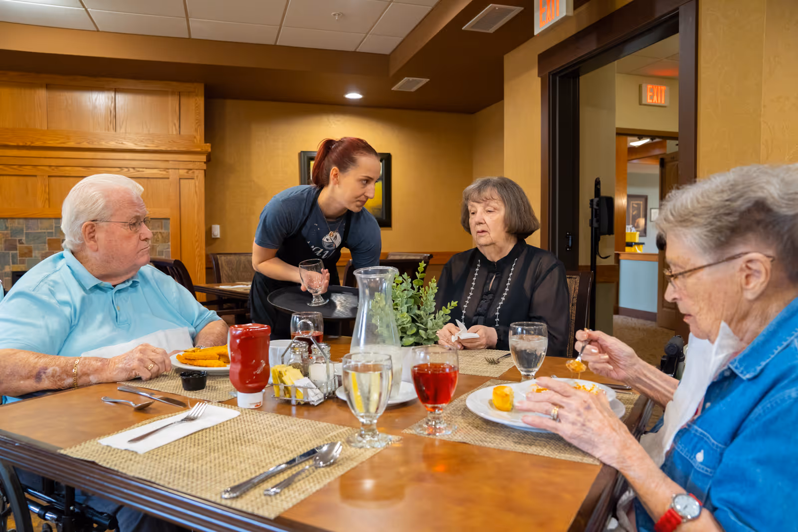 Three elderly individuals seated at a dining table in a senior living facility, with a caregiver standing and interacting with them. The table is set with plates of food, glasses of water and juice, and condiments. The room has warm lighting and wooden decor.