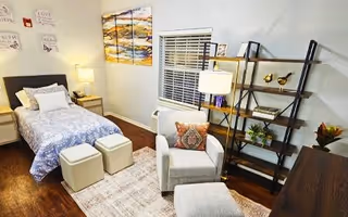 A cozy bedroom featuring a single bed with patterned bedding, two small ottomans at the foot of the bed, a gray armchair with a decorative pillow, a matching ottoman, a tall black shelving unit with decorative items, a window with white blinds, a colorful abstract painting on the wall, and a nightstand with a lamp.