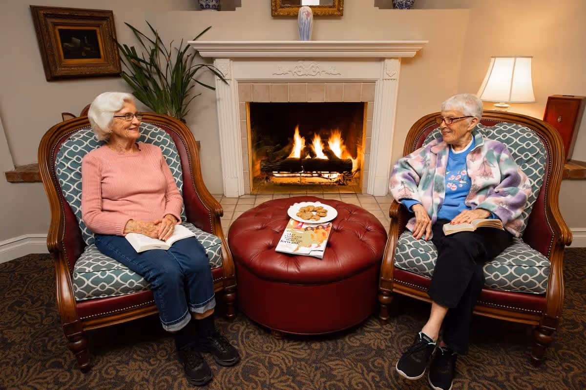Two elderly women sitting in patterned armchairs facing each other with a lit fireplace in the background. They both hold books and are smiling. Between them is a round red ottoman with a plate of cookies and some magazines on it. The room has warm lighting and a cozy atmosphere.