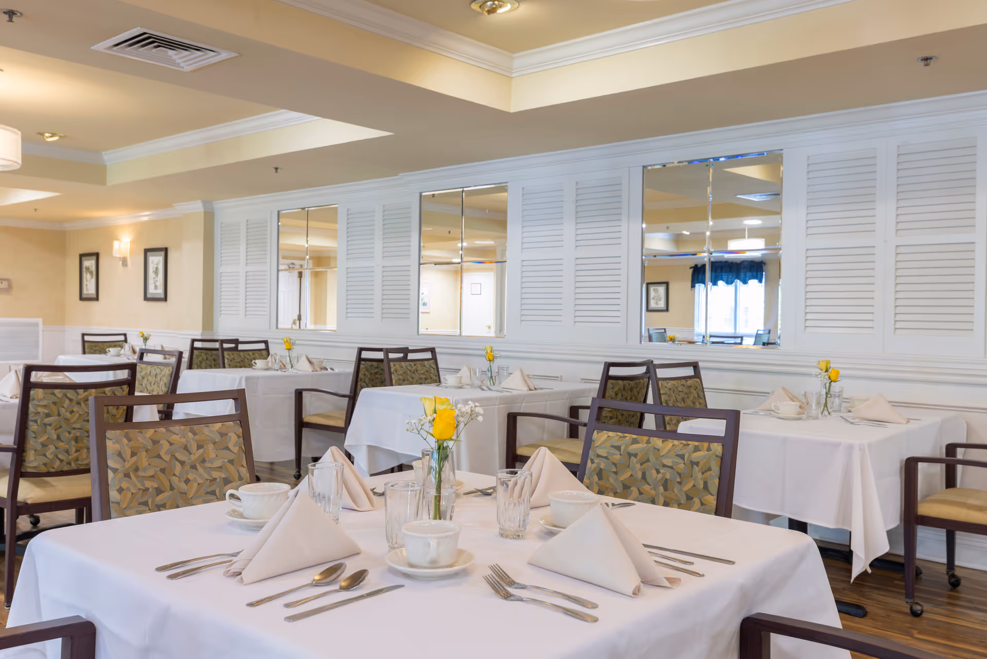 A bright and neatly arranged dining room with tables covered in white tablecloths, each set with folded napkins, cups, glasses, and silverware. Chairs with patterned upholstery surround the tables. The walls are light-colored with white paneling and mirrors, and there are framed pictures and soft lighting fixtures on the walls.
