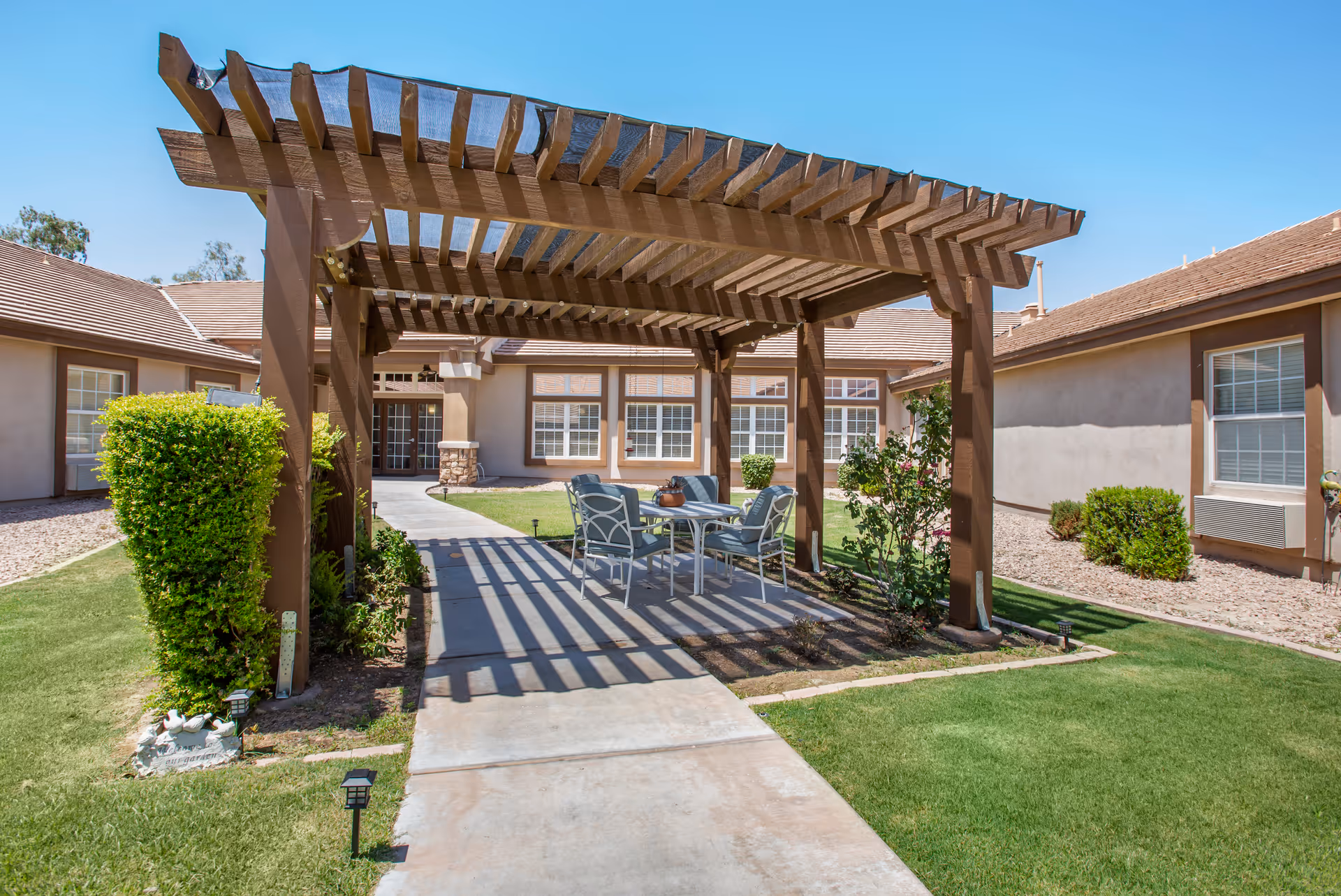 Outdoor patio area at Brookdale Chandler Ray Road featuring a wooden pergola with a mesh cover, a concrete pathway, and a table with four cushioned chairs. Surrounding the patio are well-maintained green lawns, bushes, and a beige building with multiple windows under a clear blue sky.