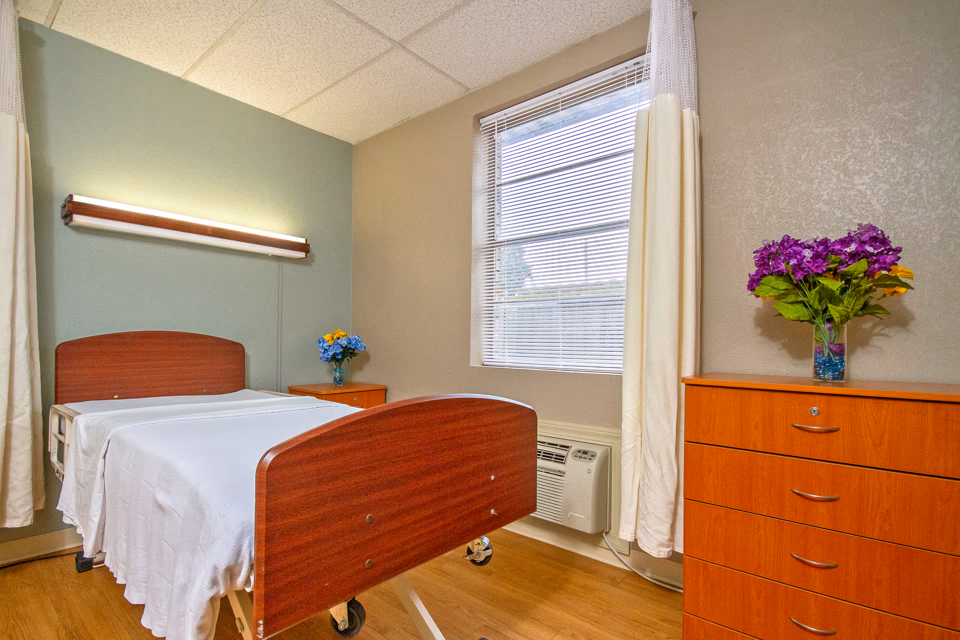 Hospital-style single bed with wooden head and footboards in a senior facility room with a dresser, nightstand, window, and vases of flowers.