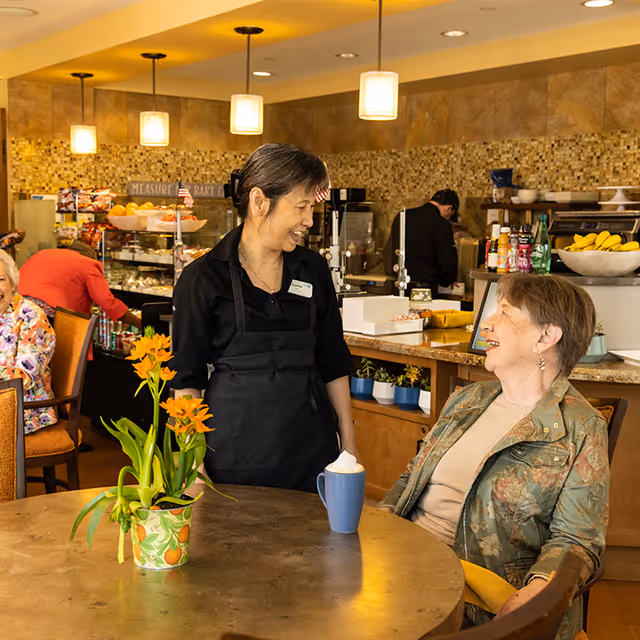 A server in a black apron smiles and talks with an elderly woman seated at a table in a bright dining area.