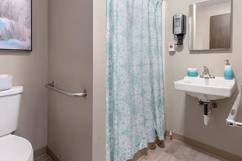 A clean and accessible bathroom featuring a toilet with a white basket on top, a wall-mounted grab bar, a shower area with a blue and white patterned curtain, a small white sink with a soap dispenser and a blue cup, a wall-mounted mirror, and a hand sanitizer dispenser.