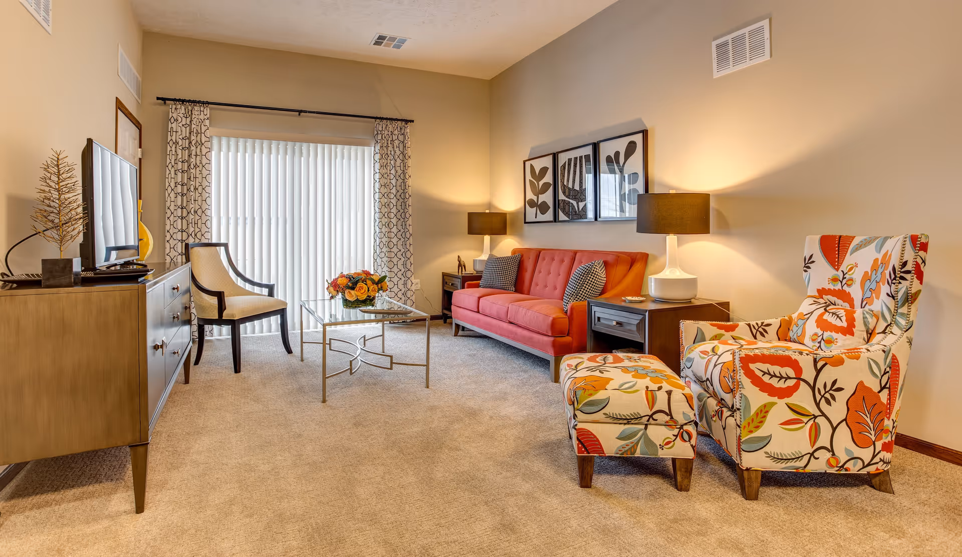 A cozy living room with a red sofa, a floral patterned armchair with matching ottoman, a glass coffee table with a flower arrangement, a wooden TV stand with a flat screen TV, and a beige armchair near a large window with vertical blinds and patterned curtains. The room is warmly lit with two table lamps and decorated with three framed botanical prints on the wall.