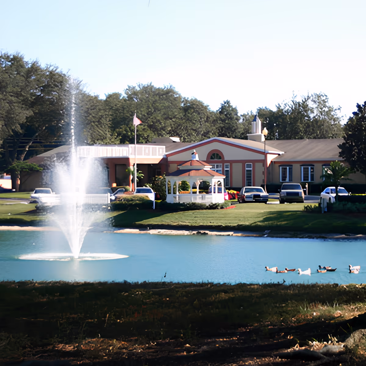 A scenic view of a senior living facility with a large pond featuring a water fountain in the foreground. There is a white gazebo near the pond, several parked cars, and a building with a peaked roof and multiple windows in the background, surrounded by trees and greenery.