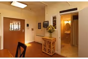 Interior view of a room with a wooden door, a small wooden table with a vase of sunflowers, a wall-mounted television, and an open doorway leading to a bathroom with a toilet and shower curtain visible.