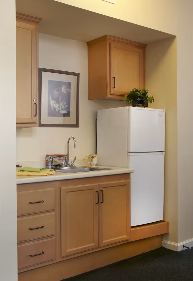 Small kitchen area with light wood cabinets, a white refrigerator, a countertop with a sink, a faucet, a yellow mug, a green towel, and a framed picture on the wall.