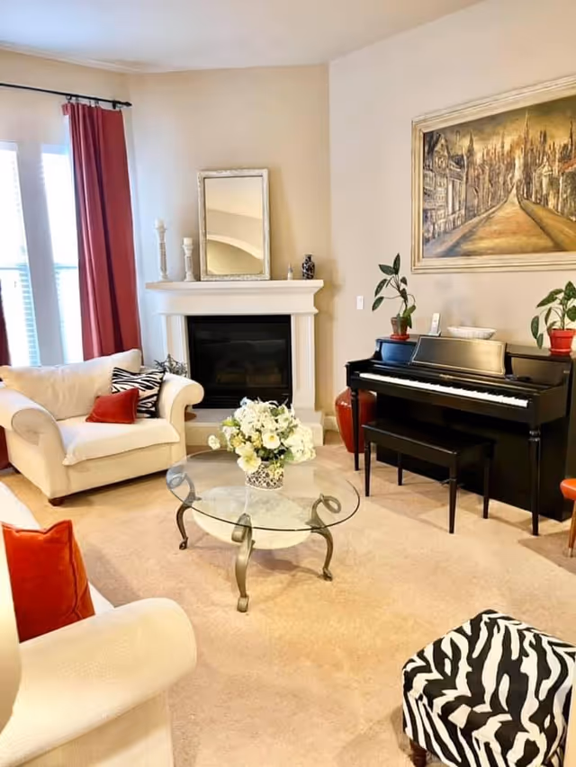 A cozy living room with beige walls and carpet, featuring a white armchair with red and zebra-patterned pillows, a glass coffee table with a floral arrangement, a black upright piano with plants on top, a fireplace with decorative items and a mirror above it, red curtains on the windows, and a large framed painting on the wall.