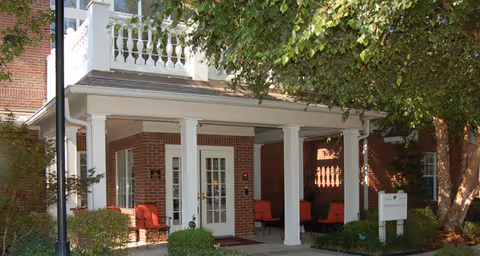 Entrance of a brick building with white columns and a covered porch area. There are red cushioned chairs arranged on the porch, surrounded by green bushes and trees providing shade.