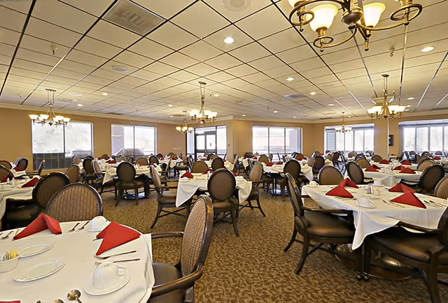 Interior view of the dining room at Pueblo Norte Senior Living, featuring elegantly set tables with white tablecloths and red napkins, surrounded by comfortable seating and chandeliers.