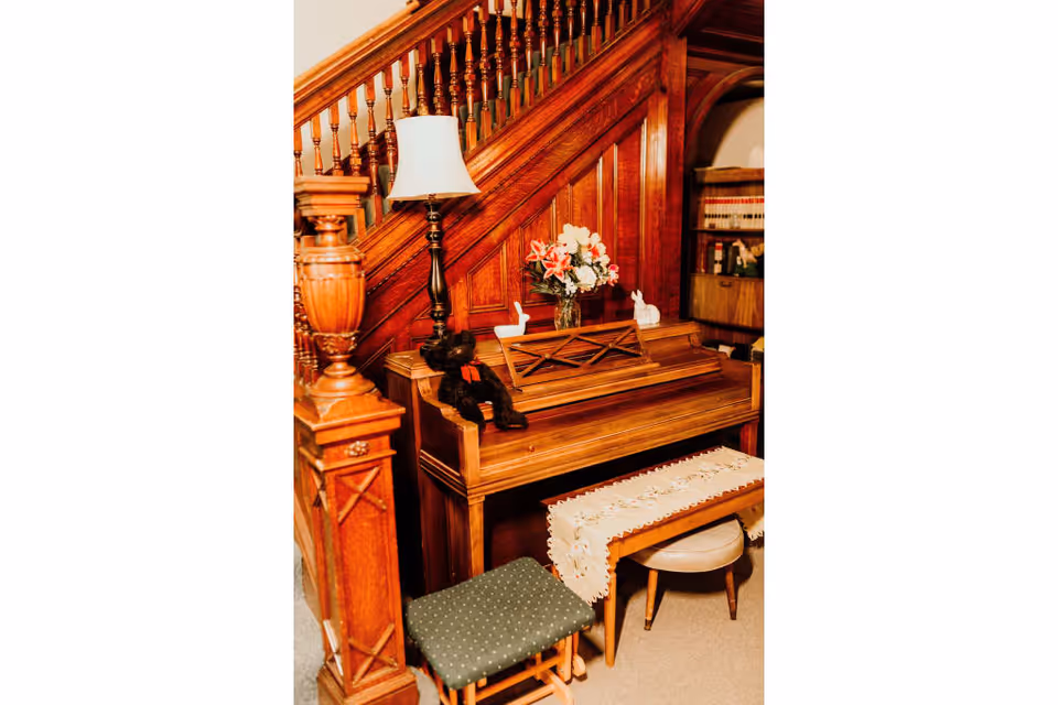 A wooden upright piano placed against a wooden staircase with ornate banisters. On top of the piano are a table lamp, a vase with flowers, and two small decorative white figurines. In front of the piano is a bench covered with a white lace runner and a small green cushioned stool. A bookshelf with books is partially visible in the background.