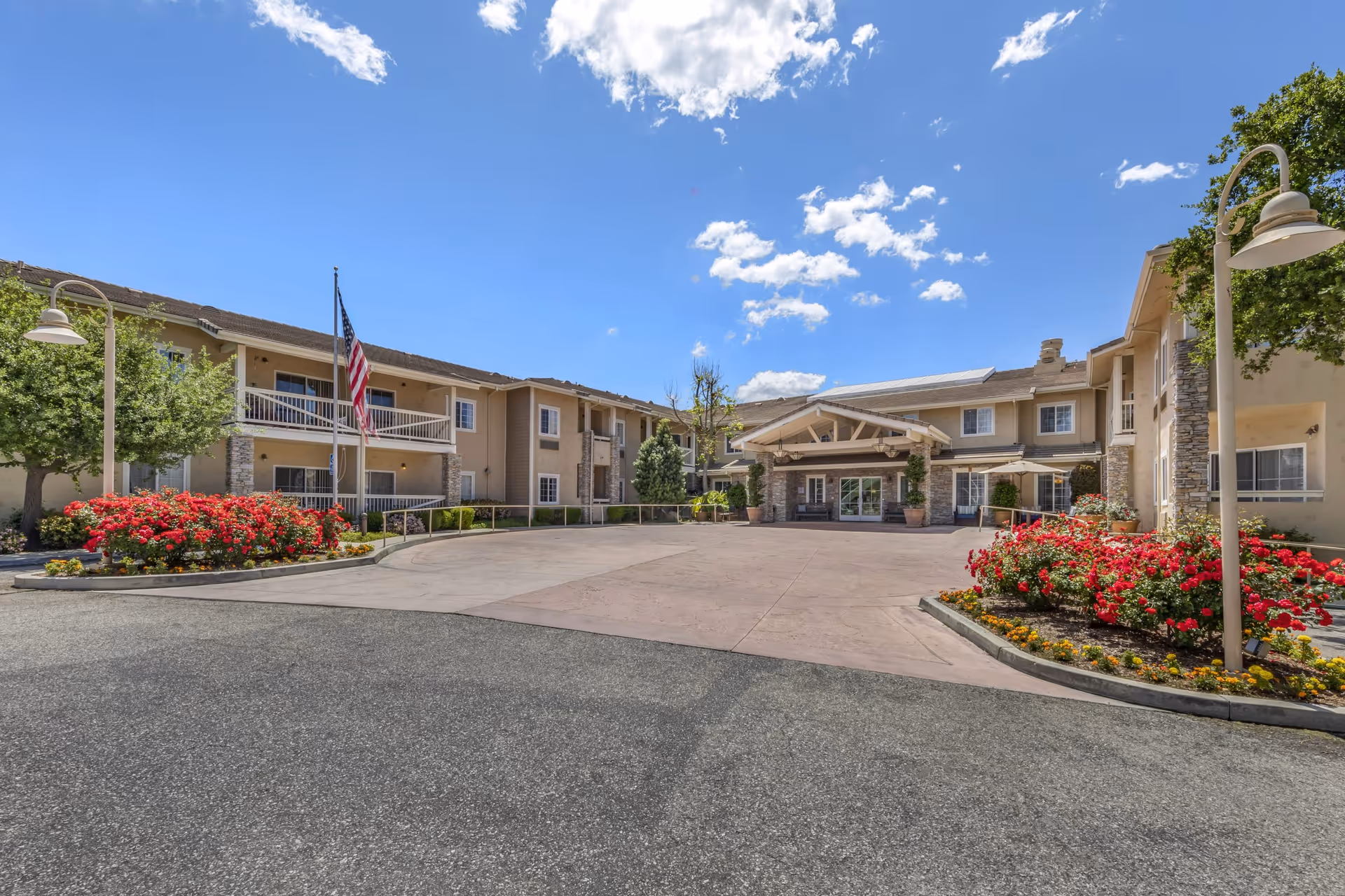 Exterior view of Meadowbrook At Agoura Hills senior living facility showing a two-story building with beige walls, stone accents, and a covered entrance. The driveway is surrounded by landscaped flower beds with red and yellow flowers, trees, and lamp posts. An American flag is displayed near the entrance under a bright blue sky with scattered clouds.