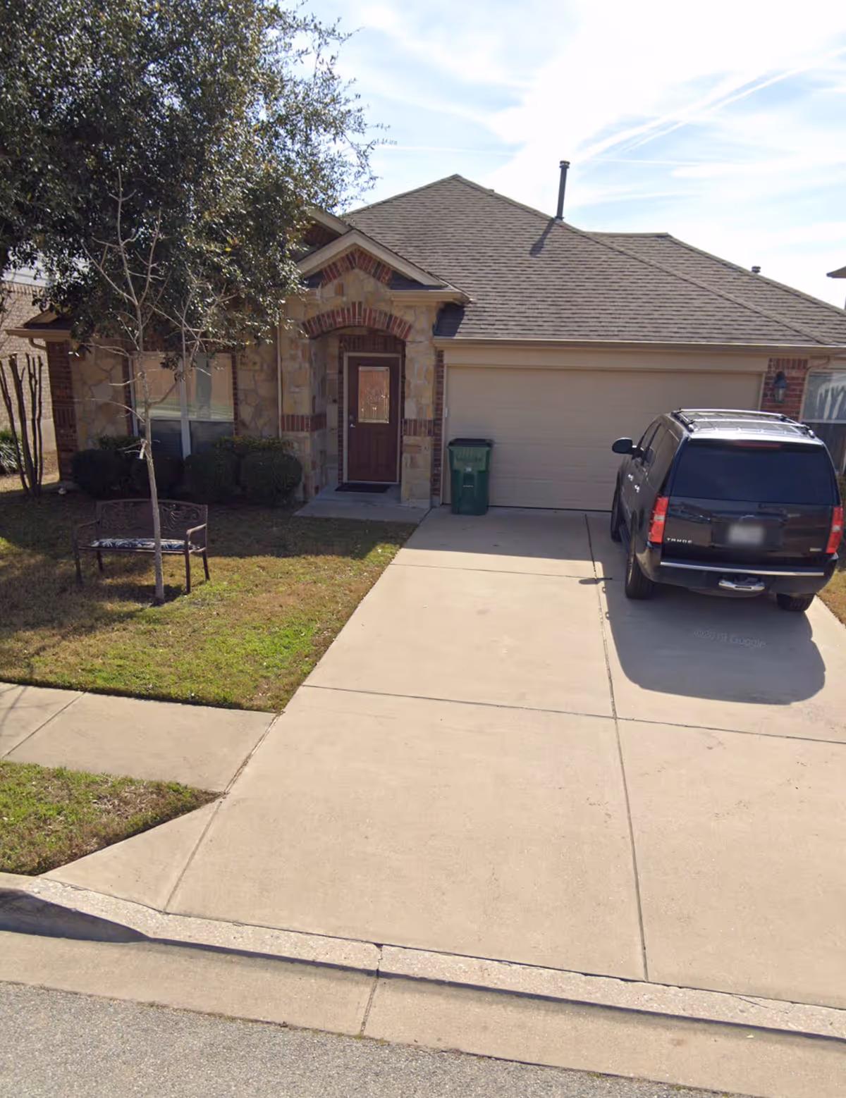 Single-story brick and stone house front with a two-car garage, a black SUV parked in the driveway, and a small lawn with a bench and tree.