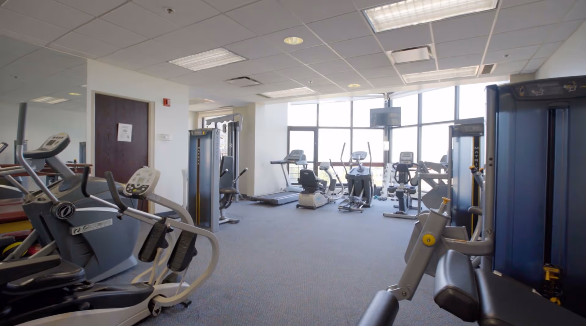 A fitness room with various exercise equipment including treadmills, stationary bikes, elliptical machines, and weight machines. The room has large windows letting in natural light and a wall-mounted TV. The ceiling has fluorescent lighting and the floor is carpeted.