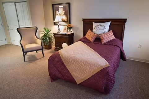 A tidy bedroom with a single bed dressed in purple and beige linens, a wooden headboard, a nightstand with a lamp and framed art, and an upholstered chair.
