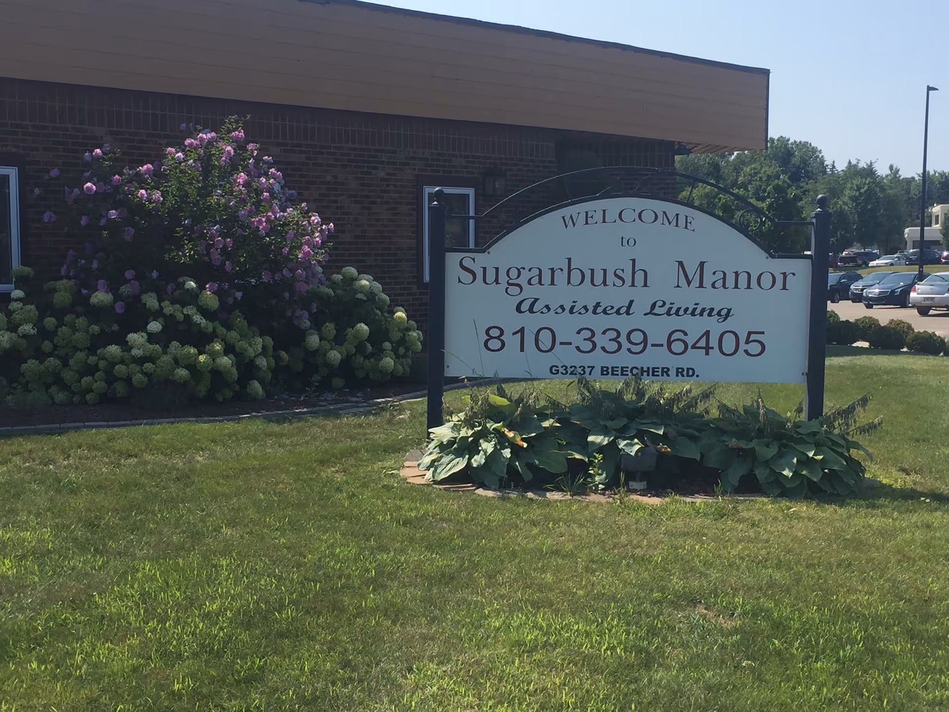 Outdoor view of a sign for Sugarbush Manor Assisted Living located on a grassy area with bushes and flowers in front of a brick building and a parking lot in the background.