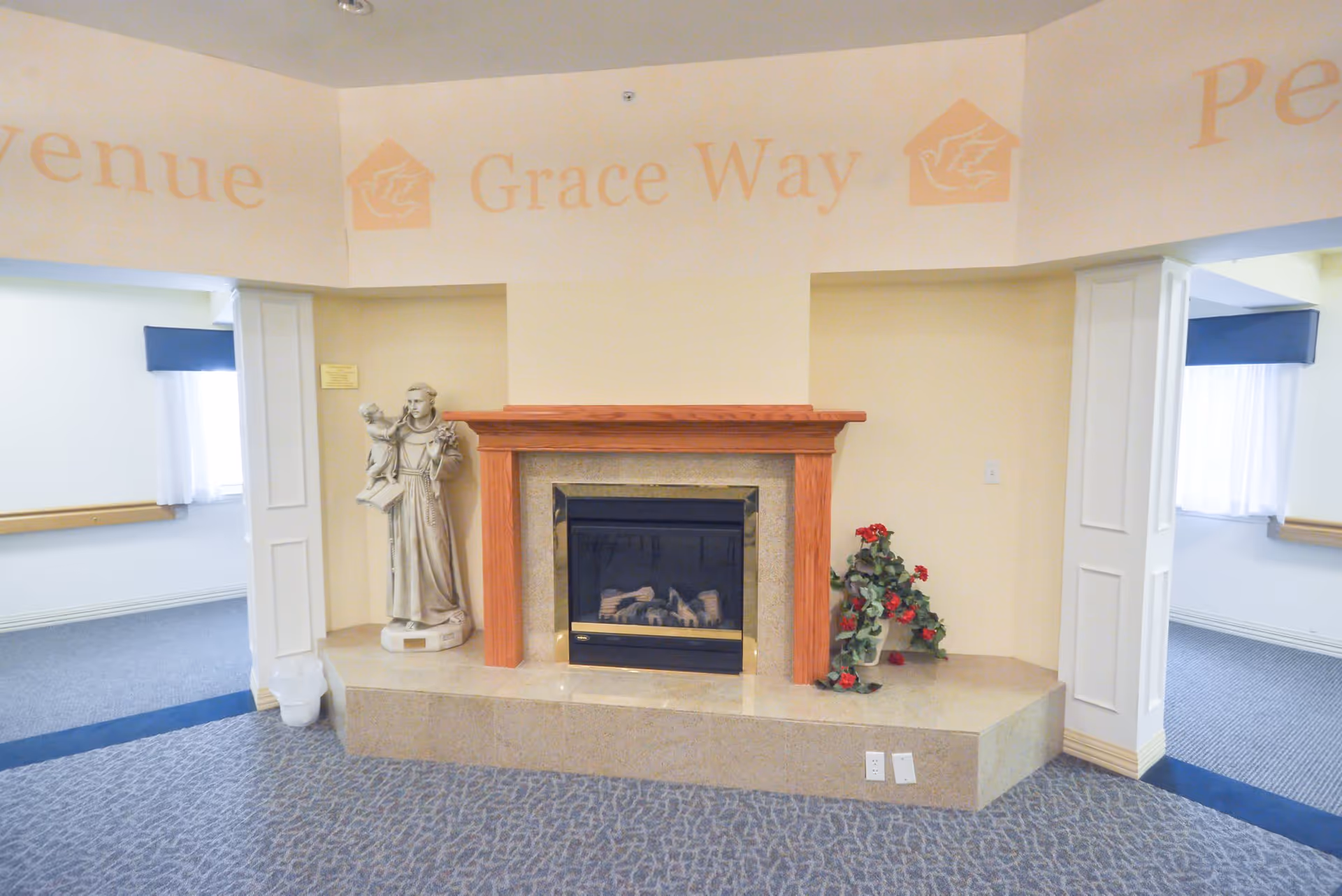 Interior view of a room with a fireplace centered on a beige wall. The fireplace has a wooden mantel and a stone base. To the left of the fireplace is a statue of a robed figure holding a child, and to the right is a plant with red flowers. The room has carpeted flooring and two open doorways on either side of the fireplace leading to adjacent rooms with windows covered by white curtains and blue valances. Above the fireplace, the words 'Grace Way' are painted on the wall.