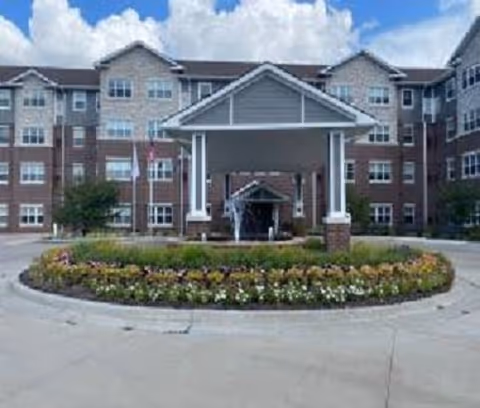 Front entrance of a multi-story senior living building with a covered porte-cochère and a circular flowerbed in the driveway.