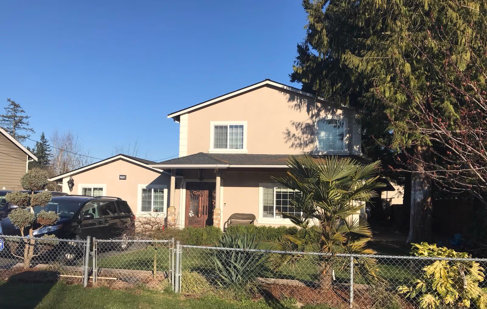Front view of a two-story beige house with a fenced yard, driveway with a parked car, and trees and shrubs in the yard.