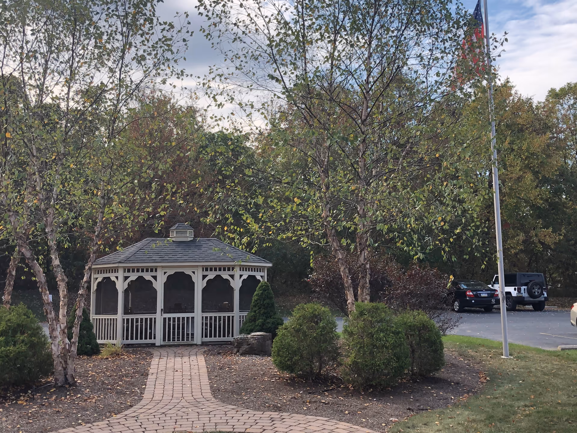A white gazebo with a shingled roof is situated at the end of a brick pathway surrounded by small trees and bushes. There is a flagpole with an American flag to the right, and a parking lot with several cars is visible in the background. The scene is outdoors with a partly cloudy sky.